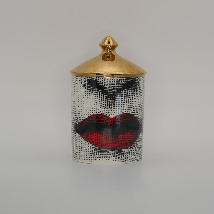 A small cylindrical container with a gold lid and a black-and-white patterned body featuring red lips and black eye makeup, centered on a light gray background.