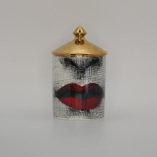 A small cylindrical container with a gold lid and a black-and-white patterned body featuring red lips and black eye makeup, centered on a light gray background.