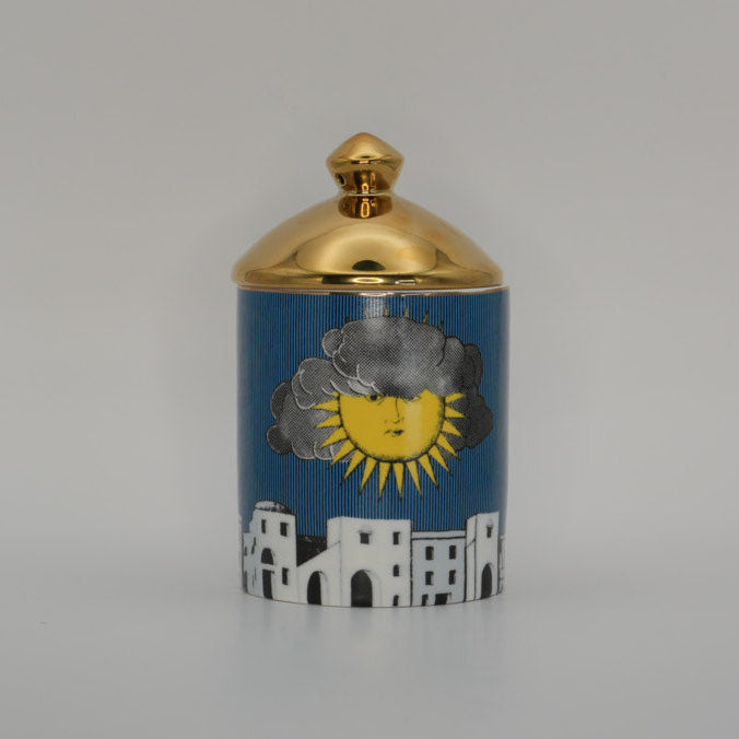 A small cylindrical lidded container with a blue illustrated body, featuring a yellow sun, gray cloud, and white building motif, topped with a shiny gold lid on a light gray background.