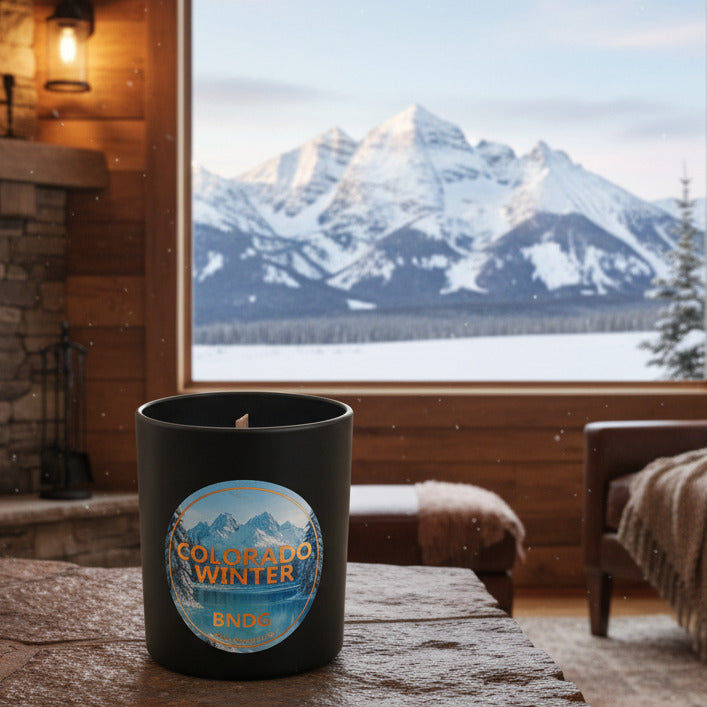 Black Candle jar with "Colorado Winter"-scent on a stone table in a cozy cabin with a view of the snowy Maroon Bells