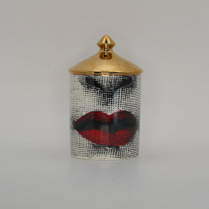 A small cylindrical container with a gold lid and a black-and-white patterned body featuring red lips and black eye makeup, centered on a light gray background.