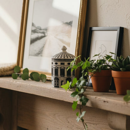 A small cylindrical lidded box or container with a detailed architectural building design in black and white, sitting on a mantle.