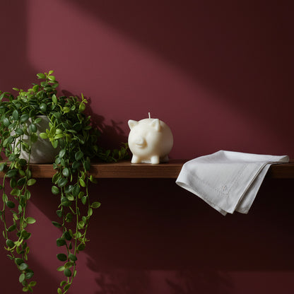 a white pig candle on a shelf with a flower and a white handkerchief with a dark red wall as background