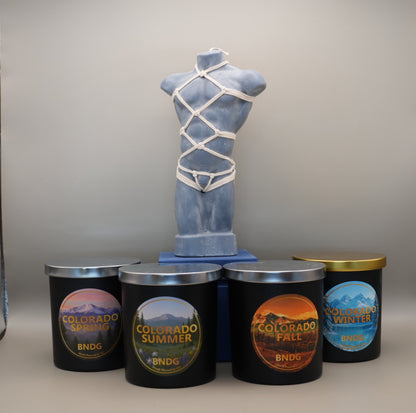 Grey Large Male Torso candle with hand-tied shibari harness, surrounded by four 12oz candle jars labeled 'Colorado Spring', 'Colorado Summer', 'Colorado Fall', and 'Colorado Winter'.